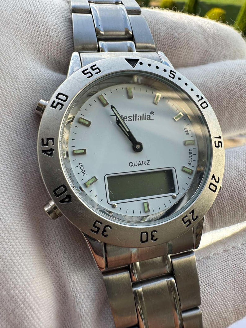 Op de afbeelding: Een zilverkleurig analoog en digitaal polshorloge met een witte wijzerplaat, voorzien van de tekst Westfalia en QUARZ. Het horloge heeft een roestvrijstalen schakelarmband, een draaibare ring met minutenaanduiding en lichtgevende uurindexen.