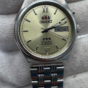 Op de afbeelding: Een zilverkleurig roestvrijstalen Orient-horloge met een champagnekleurige wijzerplaat, voorzien van een dag-datumvenster op de drie-uurpositie, Romeinse cijfers op twaalf en zes, drie sterren en de tekst Orient Crystal 21 Jewels.