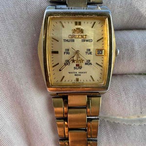 Könnte beinhalten: Goldfarbene Orient-Armbanduhr mit rechteckigem Zifferblatt. Das Zifferblatt zeigt den Wochentag, das Datum und die Uhrzeit an. Die Uhr hat ein goldfarbenes Metallarmband und ist wasserdicht bis 50m.