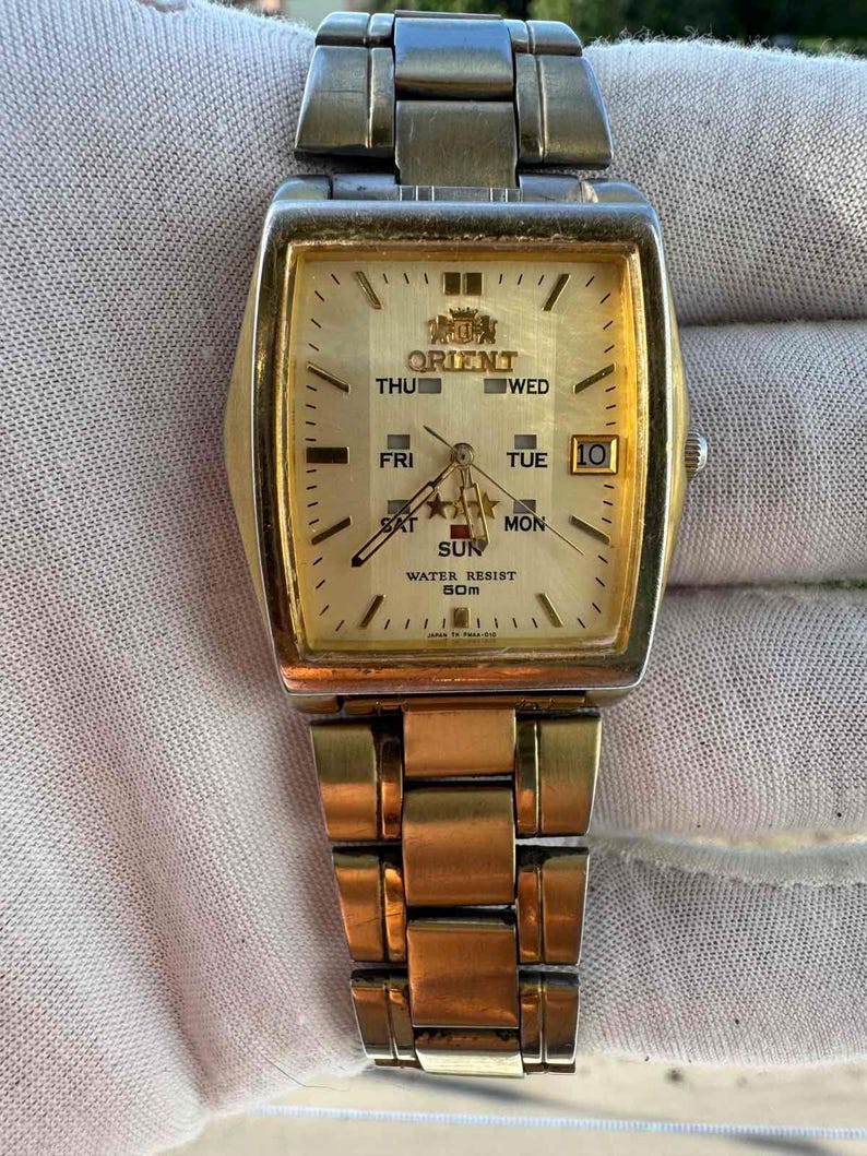 May include: A gold-toned Orient wristwatch with a rectangular face. The watch face displays the day of the week, date, and time. The watch has a gold-colored metal band and a water resistance of 50m.