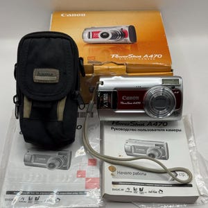 May include: A silver and maroon Canon PowerShot A470 digital camera with a wrist strap, a black Hama carrying case, an orange product box, and two instructional manuals. The camera features a lens, flash, and the text PowerShot A470.