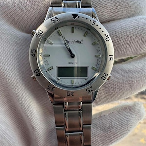 Op de afbeelding: Een zilverkleurig analoog en digitaal polshorloge met een witte wijzerplaat, voorzien van de merknaam Westfalia, het woord QUARZ en een draaibare ring met numerieke markeringen. Het horloge heeft een metalen schakelarmband en knoppen voor instellingen.