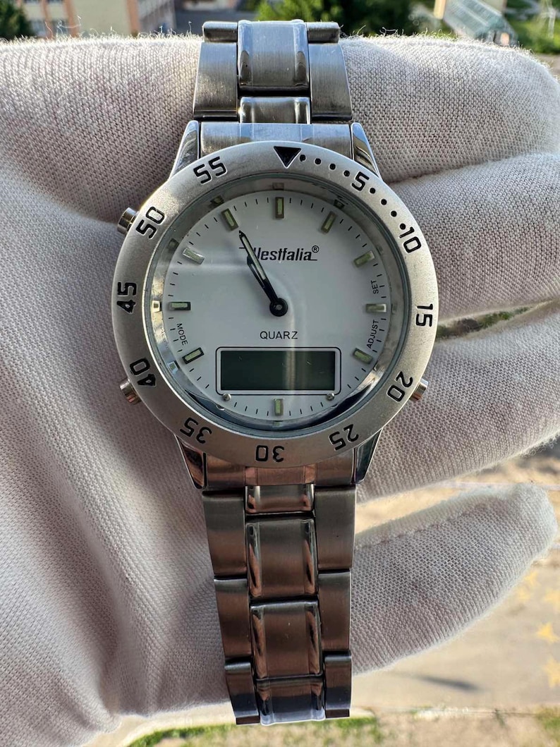 Op de afbeelding: Een zilverkleurig analoog en digitaal polshorloge met een witte wijzerplaat, voorzien van de merknaam Westfalia, het woord QUARZ en een draaibare ring met numerieke markeringen. Het horloge heeft een metalen schakelarmband en knoppen voor instellingen.