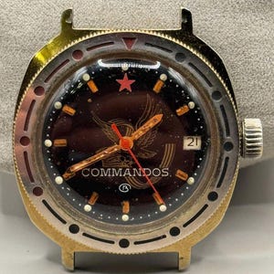 May include: A vintage wristwatch with a dark brown face featuring a red star, a gold eagle emblem, and the word "COMMANDOS." The watch has a gold-coloured bezel and a silver crown. The date is displayed at the 3 o'clock position.