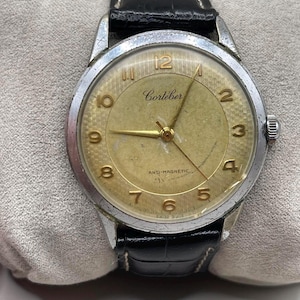 May include: Vintage Cortébert wristwatch with a gold-toned face, silver-colored bezel, and black leather band. The watch face features gold numerals and hands, with the words "Cortébert" and "ANTI-MAGNETIC" visible.
