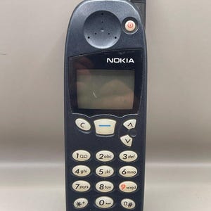 Könnte beinhalten: Ein dunkelblaues Nokia-Mobiltelefon mit kleinem Bildschirm und Tastatur. Das Telefon hat eine schwarze Antenne und der Markenname "NOKIA" ist auf der Vorderseite aufgedruckt. Das Telefon hat auch die Logos "CA'TEL" und "AT&T".