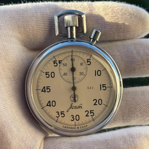 Può includere: Un cronometro meccanico color argento con quadrante color crema, numeri neri e un sotto-quadrante. Il quadrante riporta la scritta "Agat" in cirillico, "0,2 s" e "СДЕЛАНО В СССР" in basso. Include una corona superiore e un pulsante laterale.