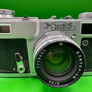 Puede incluir: Una cámara Kiev vintage plateada y negra con una lente Jupiter-8M. La cámara tiene una lente prominente, diales y la palabra "Kiev" grabada en la parte delantera. La cámara está sobre un fondo verde.