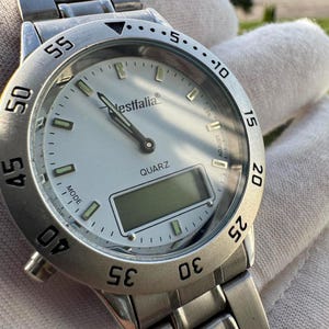 Op de afbeelding: Een zilverkleurig analoog en digitaal polshorloge met een witte wijzerplaat, voorzien van de merknaam Westfalia en het woord QUARZ op de wijzerplaat. Het horloge heeft een roestvrijstalen schakelband, een draaibare ring met minuten en lichtgevende indexen.