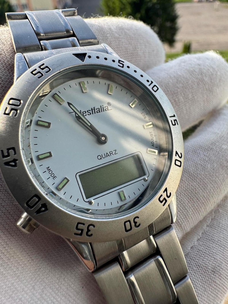 Op de afbeelding: Een zilverkleurig analoog en digitaal polshorloge met een witte wijzerplaat, voorzien van de merknaam Westfalia en het woord QUARZ op de wijzerplaat. Het horloge heeft een roestvrijstalen schakelband, een draaibare ring met minuten en lichtgevende indexen.