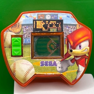 Może przedstawiać: Zabytkowa przenośna gra w baseball SEGA z Knucklesem the Echidna. Gra ma czerwoną i pomarańczową ramkę z grafiką boiska do baseballu, małym ekranem i zielonymi przyciskami sterowania. Gra pokazuje boisko do baseballu i tablicę wyników.