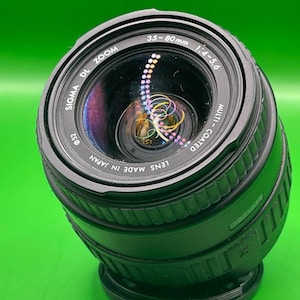 Puede incluir: Un objetivo de cámara Sigma DL Zoom 35-80mm negro con una lente multicapa. La lente tiene una rosca de filtro de 52 mm y está fabricada en Japón. La lente está sobre un fondo verde brillante.