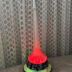 May include: A decorative lamp shaped like a rocket, featuring a silver tip and a clear, conical body that glows with red and green light. The base is black with etched details and a multi-colored, illuminated rim. The lamp is plugged into a wall outlet.
