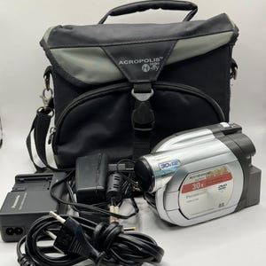 Może przedstawiać: Srebrno-czarna kamera DVD Panasonic VDR-D160 z 30-krotnym zoomem optycznym, w zestawie z czarną ładowarką Panasonic, przewodem zasilającym oraz czarno-szarą torbą transportową Acropolis Hi-fly z uchwytem i paskiem na ramię.