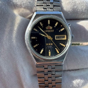 May include: A silver-tone stainless steel Orient wristwatch with a black circular dial, gold-tone hour markers, and gold-tone hands. The watch features a day-date window at the three o'clock position, three gold stars, and the text Orient Crystal 21 Jewels.
