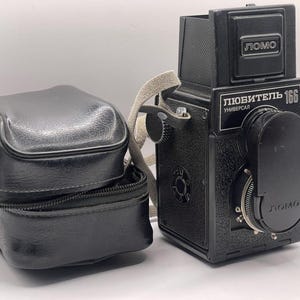 May include: A black Lomo Lubitel 166B camera with a matching black case. The camera has a lens, dials, and a strap. The case is rectangular with a zipper. The camera has the word "LOMO" on it.