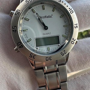 Op de afbeelding: Een zilverkleurig analoog en digitaal polshorloge met een witte wijzerplaat, voorzien van de merknaam Westfalia en het woord QUARZ op de wijzerplaat. Het horloge heeft een roestvrijstalen schakelarmband, een draaibare ring met cijfers en zijknoppen.