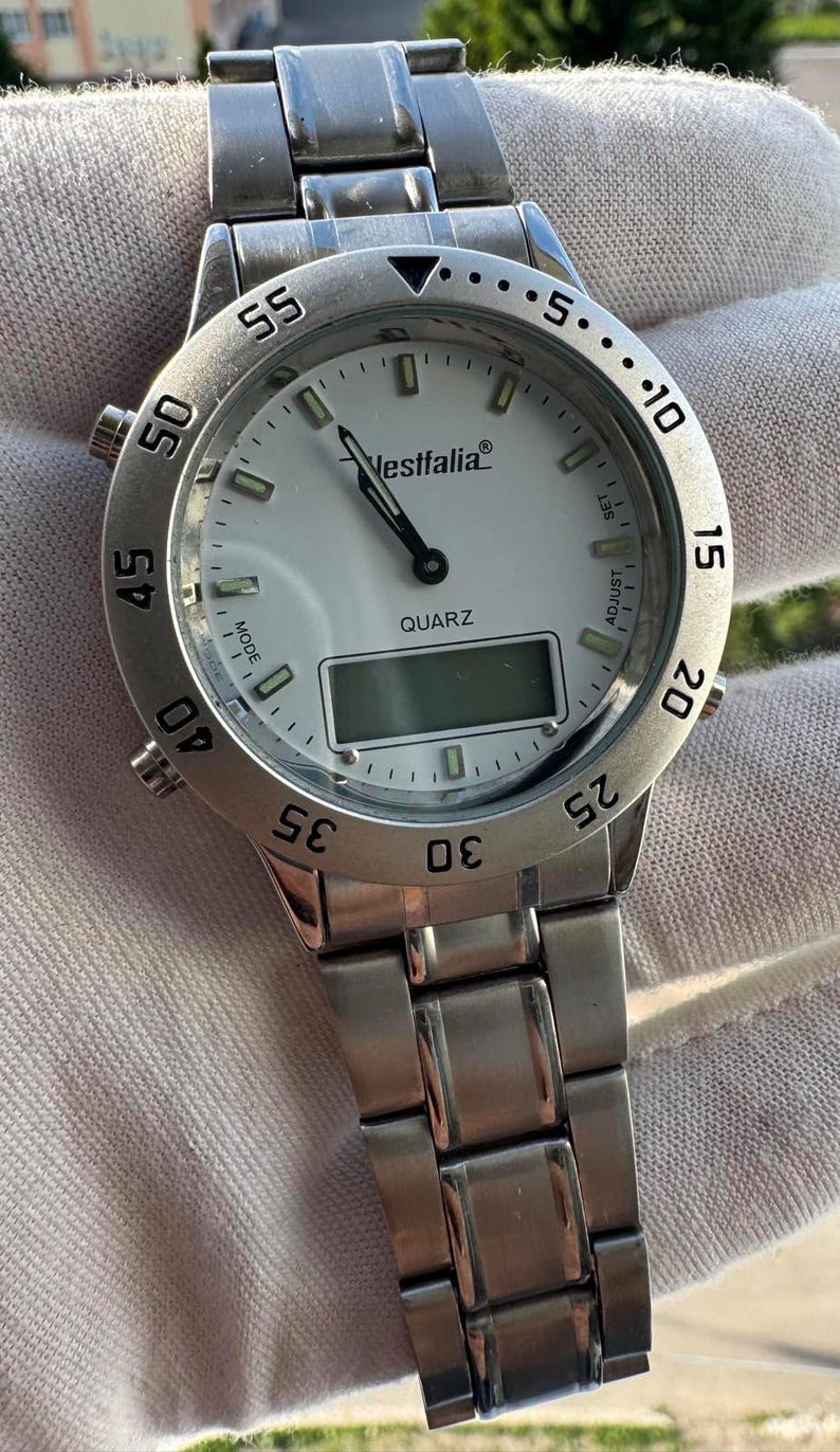 Op de afbeelding: Een zilverkleurig analoog en digitaal polshorloge met een witte wijzerplaat, voorzien van de merknaam Westfalia en het woord QUARZ op de wijzerplaat. Het horloge heeft een roestvrijstalen schakelarmband, een draaibare ring met cijfers en zijknoppen.