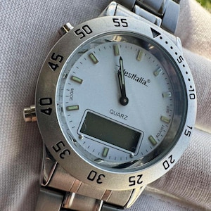 Op de afbeelding: Een zilverkleurig analoog en digitaal polshorloge met een witte wijzerplaat, voorzien van de merknaam Westfalia, het woord QUARZ en een draaibare ring met zwarte cijfers. Het horloge heeft een metalen schakelband en knoppen aan de zijkant.