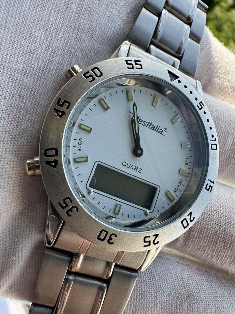 Op de afbeelding: Een zilverkleurig analoog en digitaal polshorloge met een witte wijzerplaat, voorzien van de merknaam Westfalia, het woord QUARZ en een draaibare ring met zwarte cijfers. Het horloge heeft een metalen schakelband en knoppen aan de zijkant.