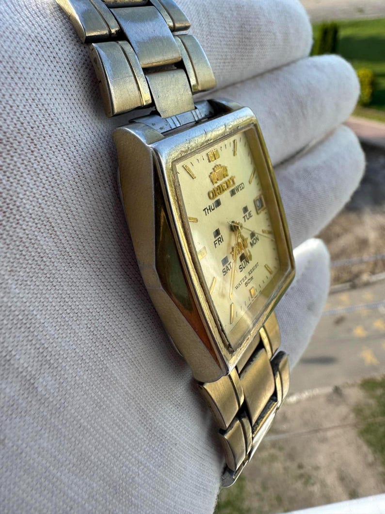 May include: A gold-toned Orient wristwatch with a rectangular face and a matching metal band. The watch face is cream-colored with gold hands and markers, and displays the day of the week. The watch is water resistant.