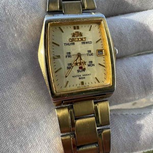 May include: A gold-toned Orient wristwatch with a rectangular face and a matching metal band. The watch face displays the day of the week and the Orient logo. The watch is water-resistant to 50m.