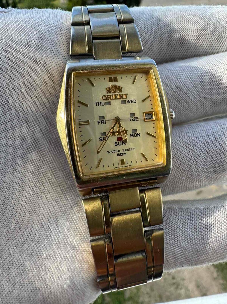 May include: A gold-toned Orient wristwatch with a rectangular face and a matching metal band. The watch face displays the day of the week and the Orient logo. The watch is water-resistant to 50m.