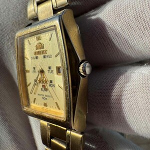 May include: A gold-toned Orient wristwatch with a rectangular face. The watch face displays the day of the week and the time. The watch band is also gold-toned and made of linked metal pieces. The watch is water resistant to 50m.