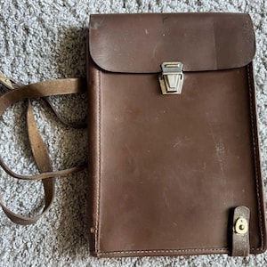 May include: A brown leather vertical messenger bag featuring a fold-over flap with a silver-toned metal clasp closure, a long adjustable shoulder strap, and a small vertical leather tab with a brass-toned snap button fastener at the bottom right.
