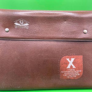 May include: Brown leather document case with a flap closure secured by two silver rivets. The case features a logo and text in Cyrillic script, along with a red and white emblem. The case is rectangular and appears vintage.