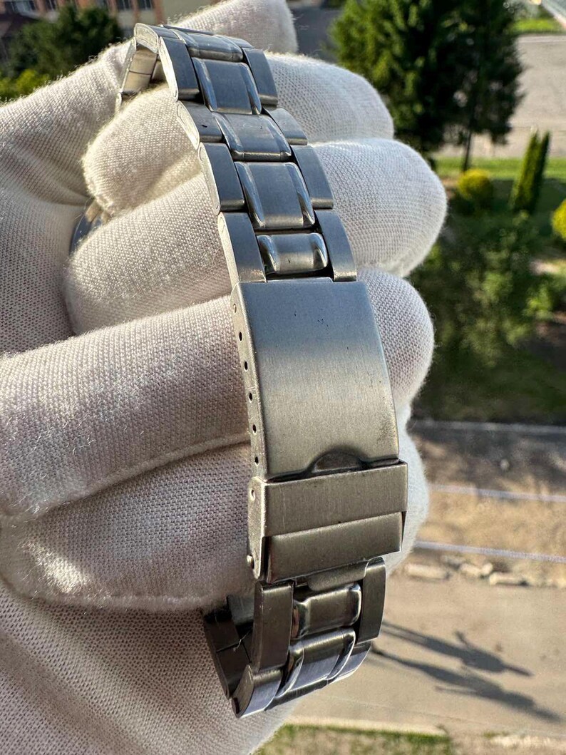 Op de afbeelding: Een zilverkleurige metalen horlogeband met een reeks rechthoekige schakels en een vouwsluiting. De band heeft een geborstelde afwerking en is opgebouwd uit meerdere in elkaar grijpende segmenten die een flexibele riem voor een horloge vormen.