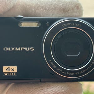 May include: A black Olympus digital camera featuring a 4x wide optical zoom lens with 4.8-19.2mm 1:2.9-6.5 specifications. The front of the camera displays the Olympus brand name, a flash unit, and a rectangular badge indicating 4x wide zoom.