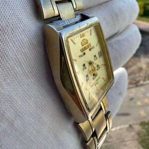 May include: A gold-toned Orient wristwatch with a rectangular face and a matching metal band. The watch face is cream-colored with gold hands and markers, and displays the day of the week. The watch is water resistant.