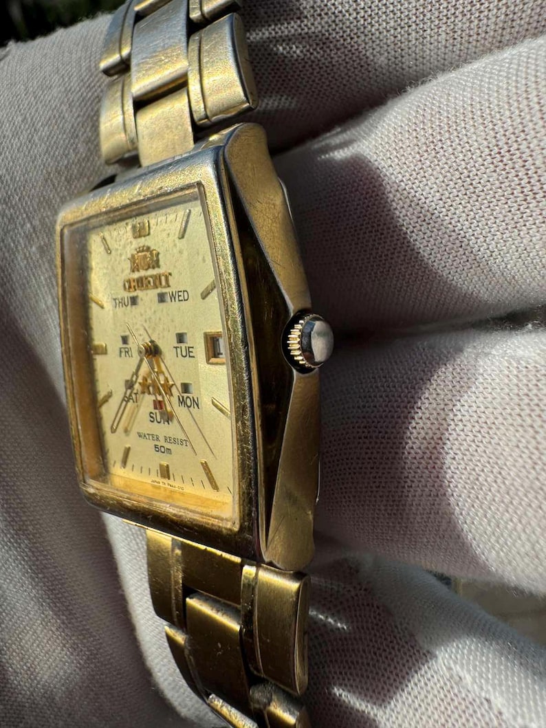 May include: A gold-toned Orient wristwatch with a rectangular face. The watch face displays the day of the week and the time. The watch band is also gold-toned and made of linked metal pieces. The watch is water resistant to 50m.