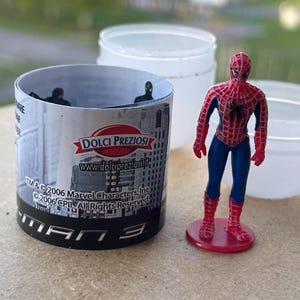 Pode incluir: Uma figura do Homem-Aranha em uma base vermelha ao lado de um recipiente cil&iacute;ndrico com um design de paisagem urbana e o texto "MAN 3". Dois recipientes de pl&aacute;stico branco ao fundo.