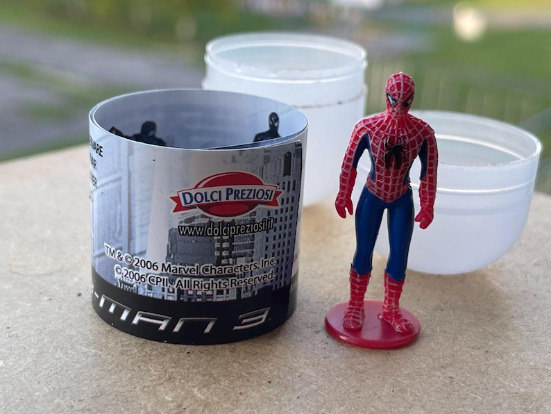 Pode incluir: Uma figura do Homem-Aranha em uma base vermelha ao lado de um recipiente cil&iacute;ndrico com um design de paisagem urbana e o texto "MAN 3". Dois recipientes de pl&aacute;stico branco ao fundo.