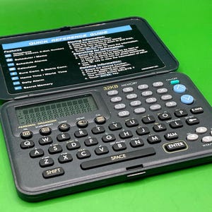 Può includere: Un'agenda elettronica nera vintage con tastiera, schermo e coperchio aperto che rivela una guida di riferimento rapida. Il dispositivo ha pulsanti e 32 KB di memoria.