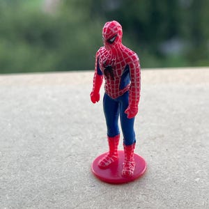 Pode incluir: Uma pequena figura do Homem-Aranha, em p&eacute; sobre uma base circular vermelha. A figura est&aacute; com o cl&aacute;ssico traje do Homem-Aranha com as cores vermelha e azul. O fundo &eacute; verde desfocado.