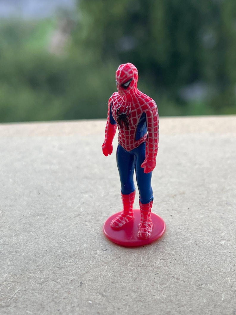 Pode incluir: Uma pequena figura do Homem-Aranha, em p&eacute; sobre uma base circular vermelha. A figura est&aacute; com o cl&aacute;ssico traje do Homem-Aranha com as cores vermelha e azul. O fundo &eacute; verde desfocado.