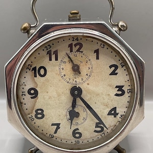 Puede incluir: Reloj despertador plateado vintage con esfera color crema y números negros. El reloj tiene un asa y dos patas pequeñas. La esfera presenta un dial más pequeño y dos manecillas que indican la hora.
