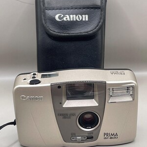 May include: A silver Canon Prima BF-800 35mm film camera with a Canon lens 28mm. The camera has a built-in flash and an on/off switch. A black leather Canon case is behind the camera.