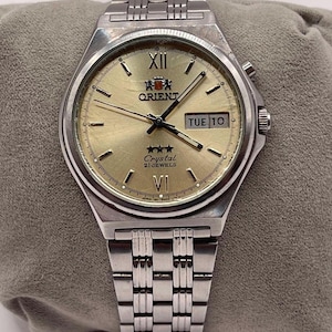 Può includere: Un orologio Orient argentato con quadrante dorato. L'orologio mostra giorno e data, con "TUE 10". Ha un cinturino argentato e un quadrante rotondo con numeri romani. Design classico.