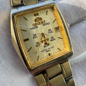 May include: A gold-toned Orient watch with a rectangular face. The watch face displays the days of the week, the date, and the Orient logo. The watch band is also gold-toned and made of linked metal pieces. The watch is water resistant to 50m.