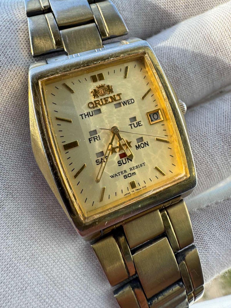 May include: A gold-toned Orient watch with a rectangular face. The watch face displays the days of the week, the date, and the Orient logo. The watch band is also gold-toned and made of linked metal pieces. The watch is water resistant to 50m.