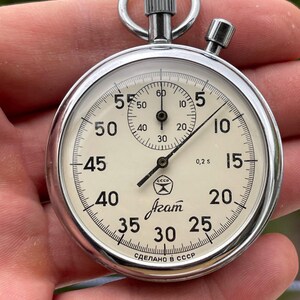 May include: A vintage stopwatch with a silver casing and a cream-colored face. The face features black numerals, a smaller dial, and the text "CCCP" and "Azam". The stopwatch is marked "Made in USSR".