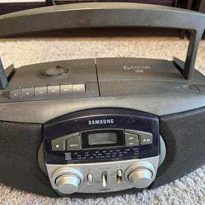 Könnte beinhalten: Ein dunkelgrauer Samsung-Boombox mit Griff, Lautsprechern und digitaler Anzeige. Das Radio verfügt über einen CD-Player und einen Tuner mit einer Reihe von Frequenzen. Die Boombox hat ein dunkelblaues Bedienfeld mit silbernen Knöpfen.