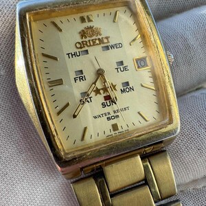 May include: A gold-toned Orient watch with a rectangular face. The watch face displays the day of the week, date, and the Orient logo. The watch band is also gold-toned. The watch is marked "WATER RESIST 50m" and "JAPAN TH PMAA-010".