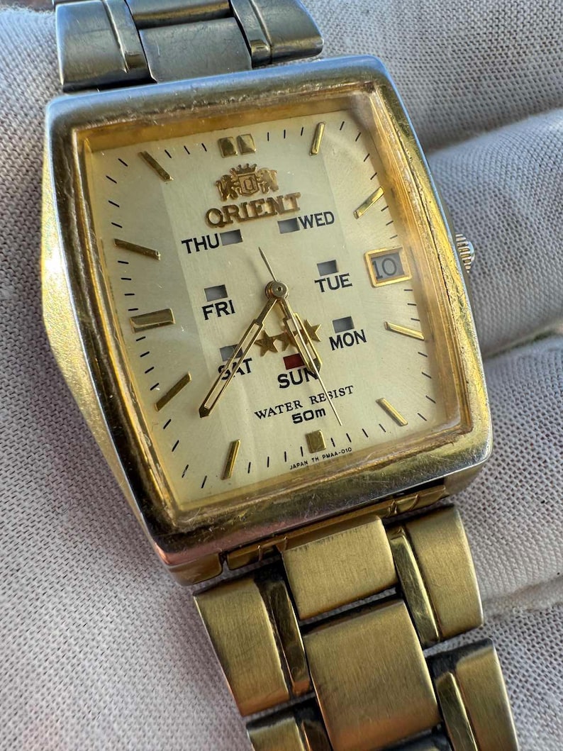 May include: A gold-toned Orient watch with a rectangular face. The watch face displays the day of the week, date, and the Orient logo. The watch band is also gold-toned. The watch is marked "WATER RESIST 50m" and "JAPAN TH PMAA-010".