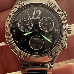 May include: A Swatch Swiss watch with a black face, silver bezel, and a stainless steel band. The watch features a chronograph function with three sub-dials, luminous hands, and the date displayed at the 3 o'clock position. The watch is approximately 40mm in diameter.