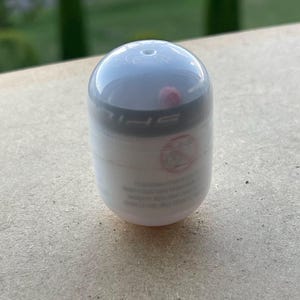Pode incluir: Um recipiente branco e cinza em forma de c&aacute;psula com uma parte superior arredondada. O recipiente tem uma faixa preta ao redor do meio e algum texto. O fundo &eacute; uma superf&iacute;cie marrom clara e folhagem verde.